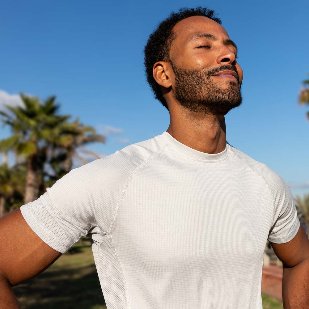 A man in a white athletic shirt stands outdoors near palm trees, breathing deeply with a calm and confident expression.