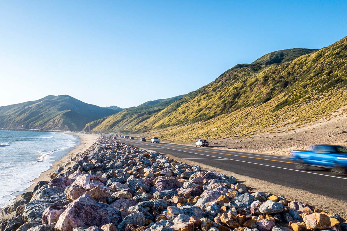 A coastal road curves along the shoreline with cars traveling beside ocean waves and rugged mountains under a bright blue sky.