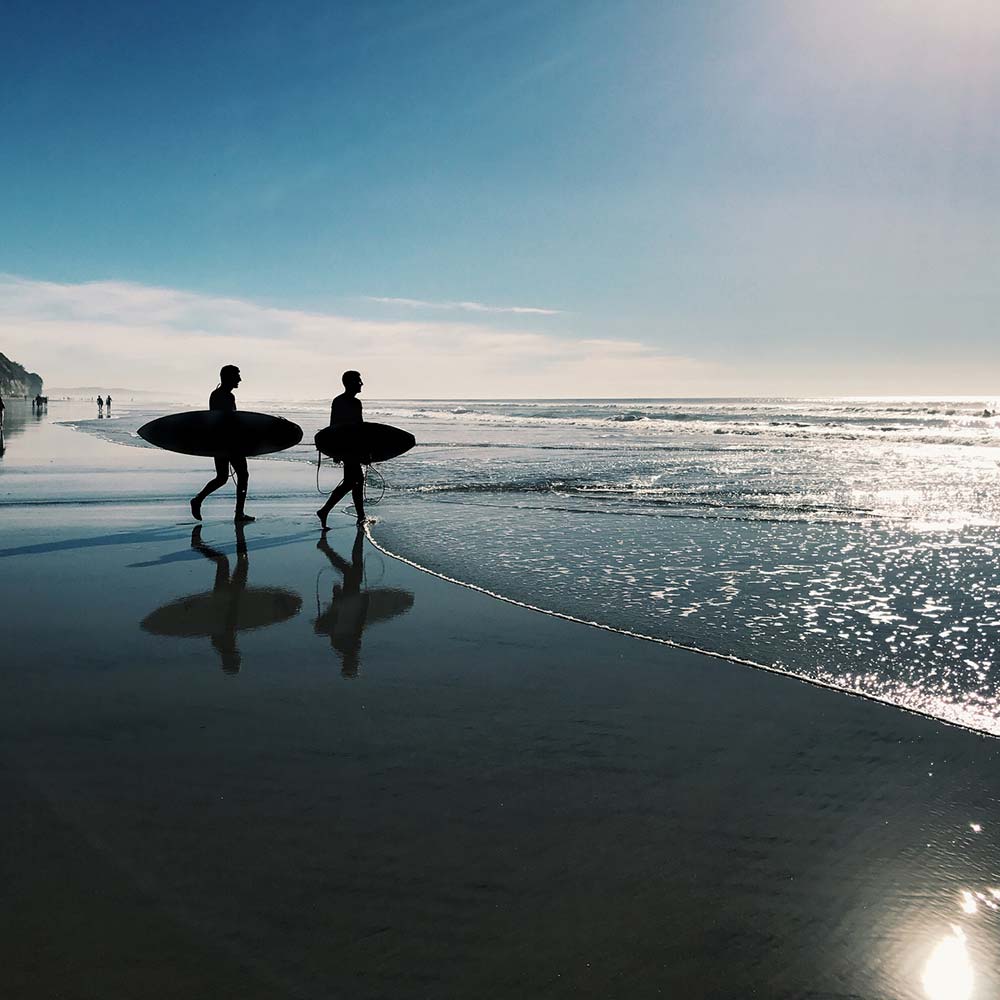 Two silhouetted surfers walk along the beach, reflecting in the wet sand as the sun glistens off the water.