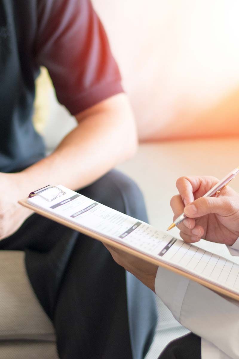 A close-up of a therapist taking notes on a clipboard while a man sits nearby with his hands clasped during a consultation.