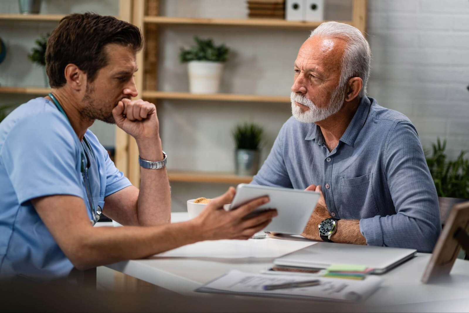 A medical professional sits across from an older man at a table, discussing information on a tablet during a clinical consultation.