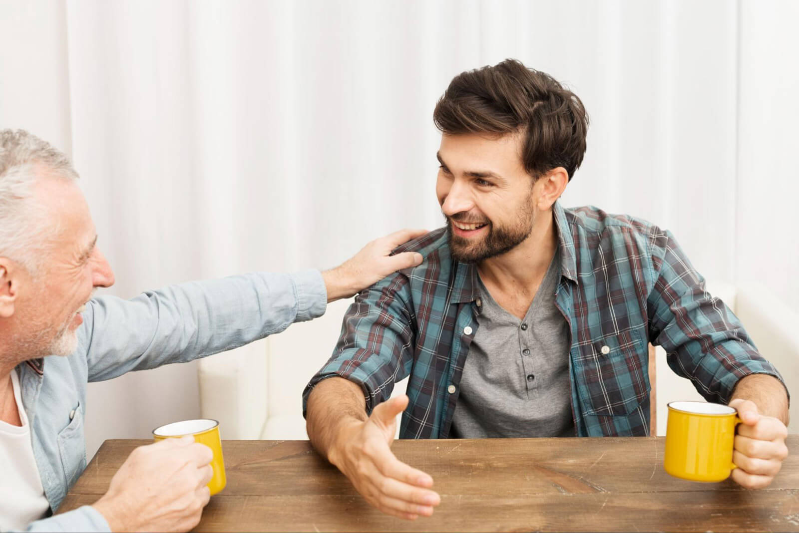 A younger man and an older man sit at a table with coffee mugs, smiling and talking in a supportive conversation.