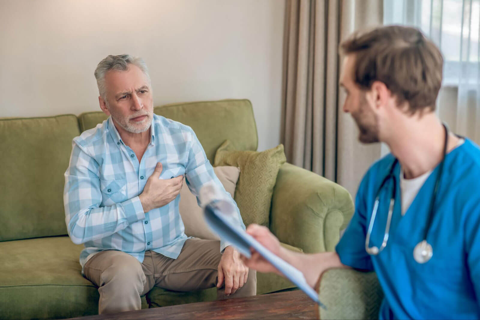A man sitting on a couch holding his chest while speaking with a healthcare professional, suggesting a medical consultation and concern about physical or emotional well-being.