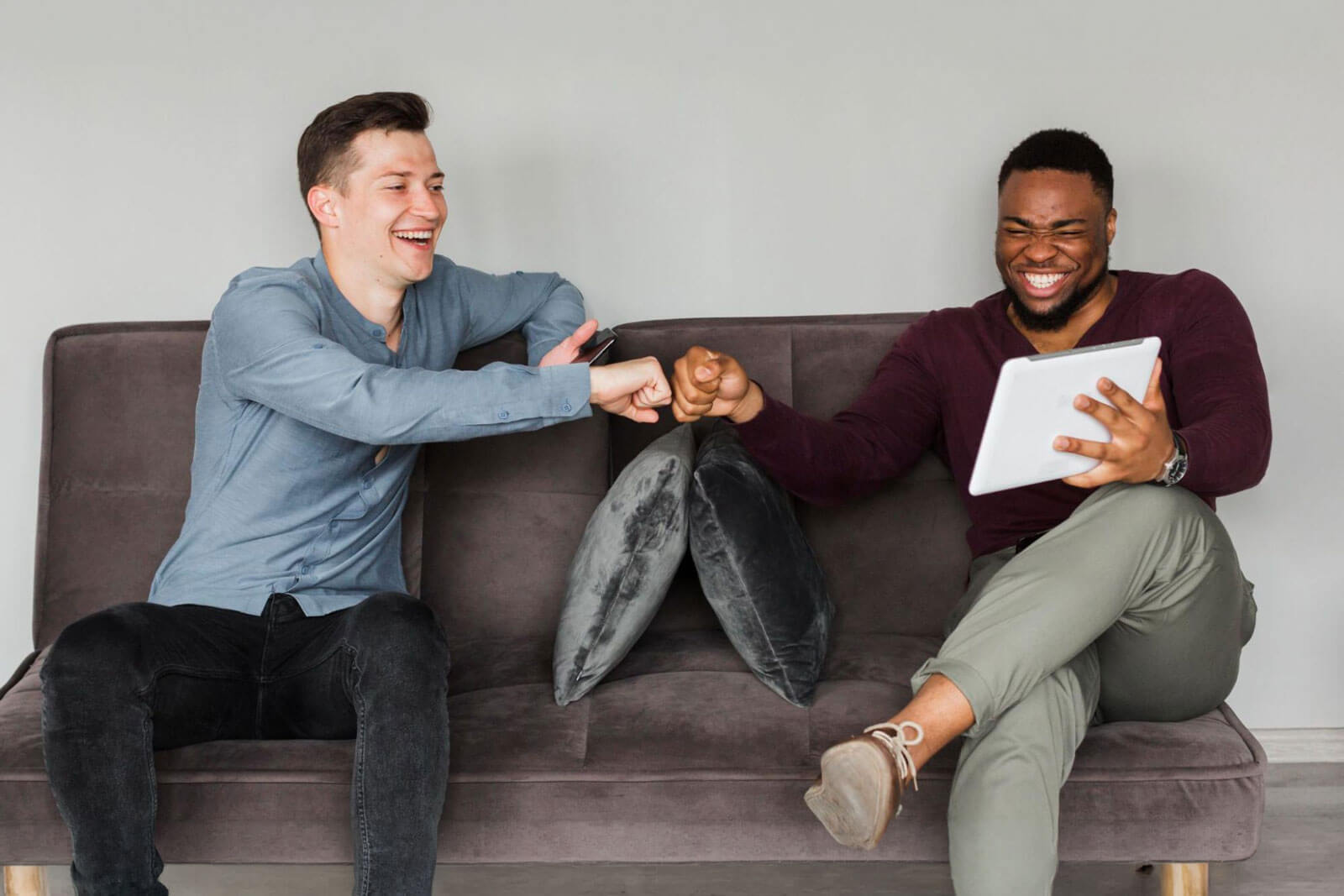 Two men — one in a blue shirt and one in a maroon sweater holding a tablet — laugh and fist-bump while sitting together on a gray sofa.
