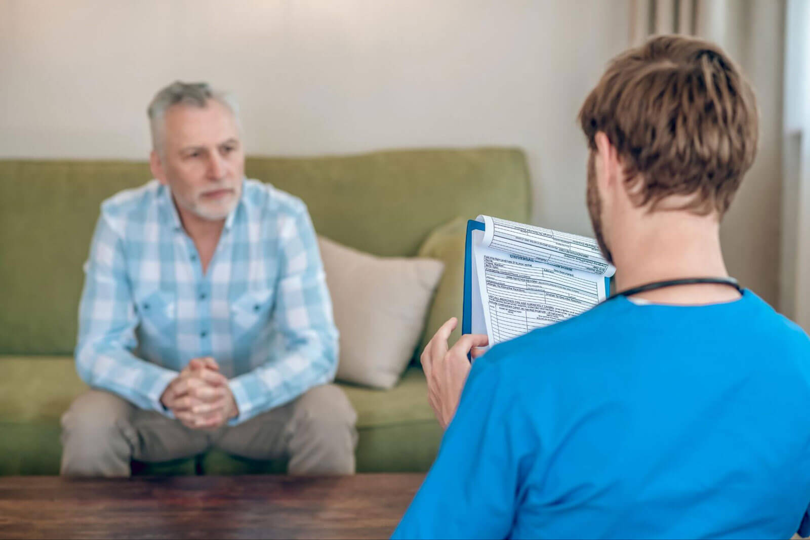 An older man with gray hair and a beard sits on a green sofa with clasped hands, listening as a healthcare provider in a blue top holds a clipboard during a home visit consultation.