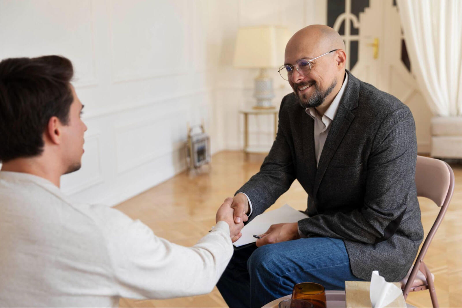 A smiling bald counselor in a gray blazer and glasses shakes hands warmly with a young male client in a bright, elegantly furnished therapy room.