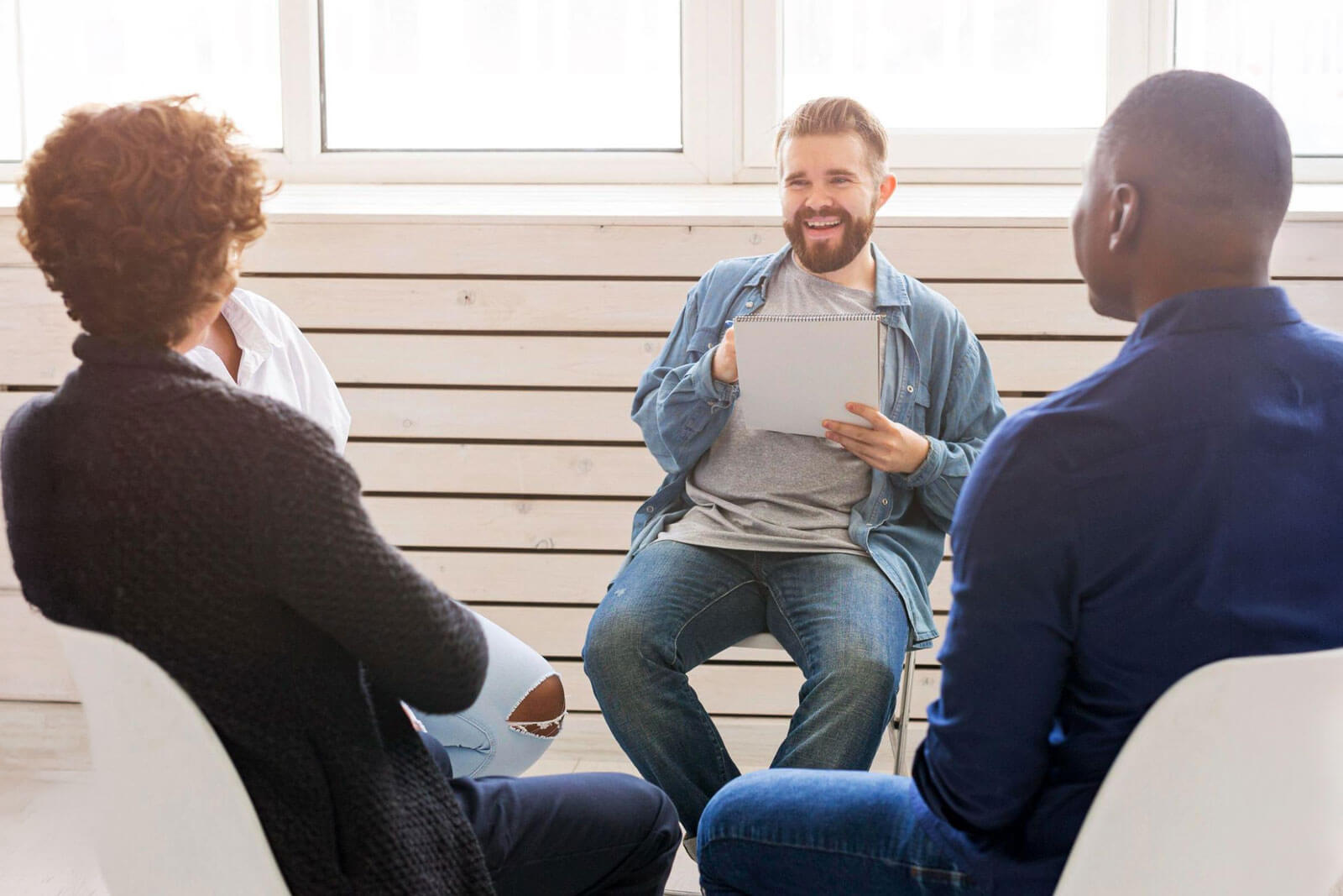 A man smiles while holding a notepad and speaking to two others during a group therapy session.