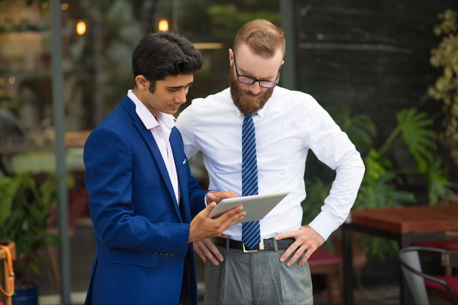 Two businessmen in suits reviewing information together on a tablet outdoors.