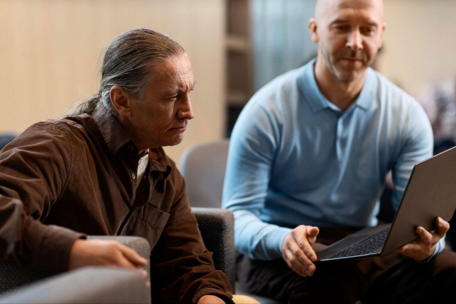 Two men sit together looking at a laptop, one with a concerned expression and the other explaining something.