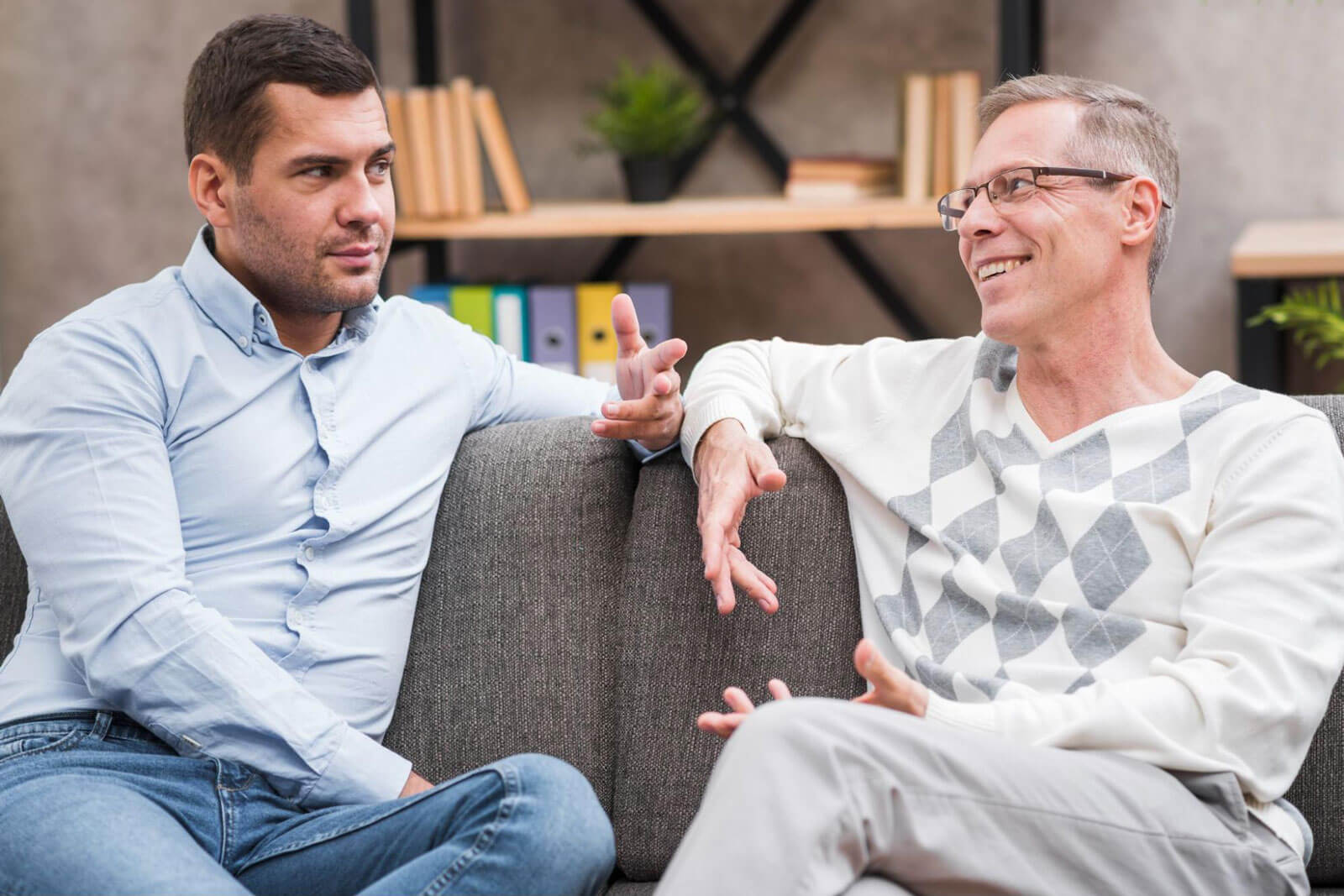 Two men sitting on a couch having a conversation with expressive hand gestures.