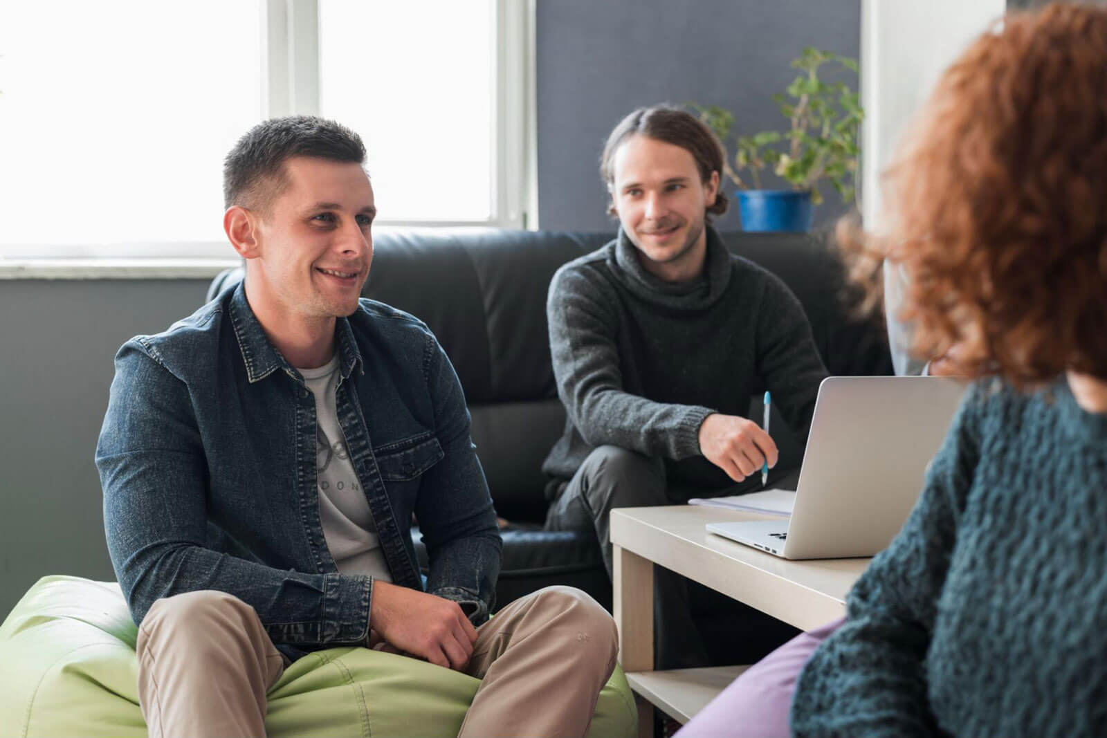Three people in a casual meeting room discussing something around a laptop on a table.