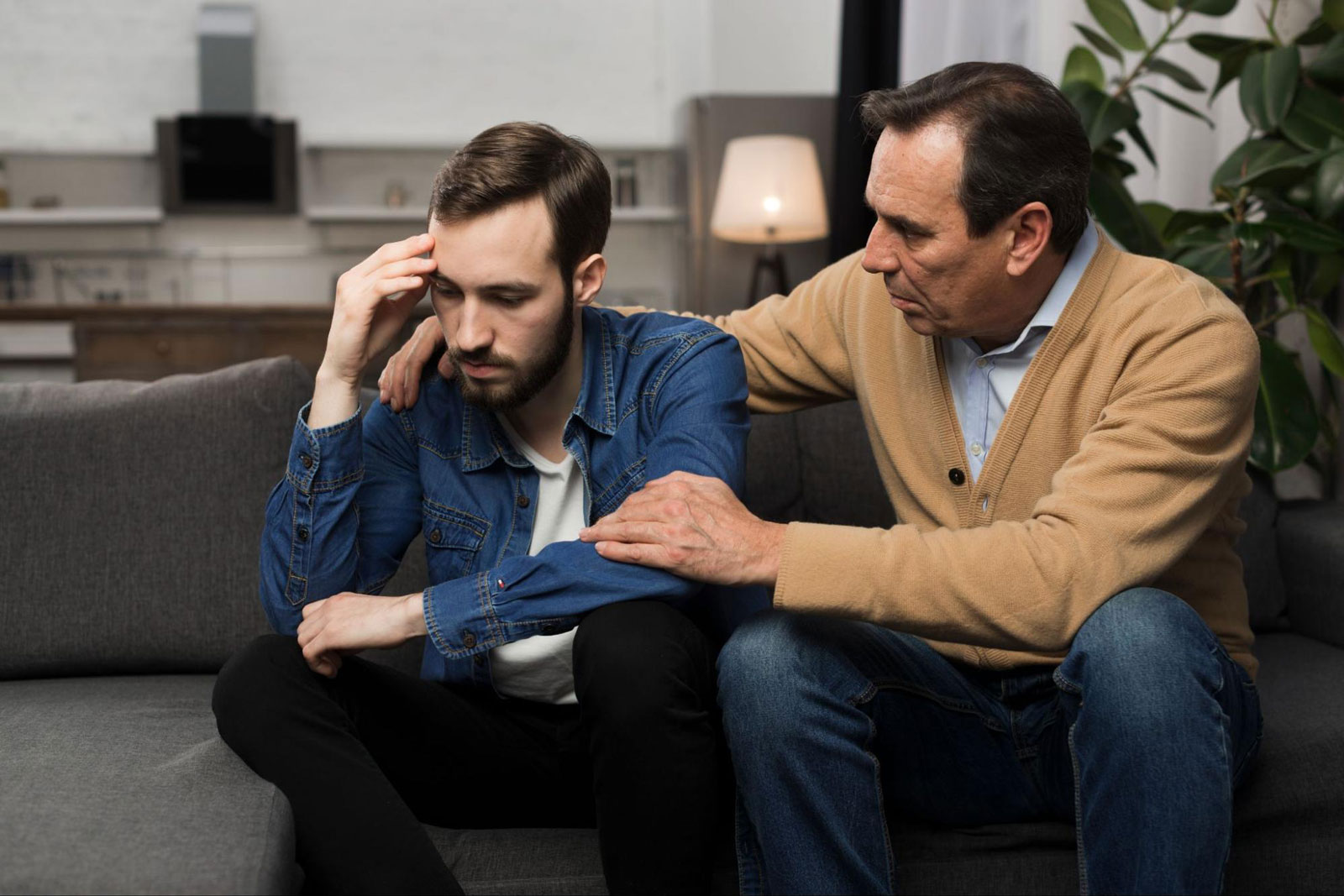 An older man comforts a distressed younger man sitting beside him on a couch.